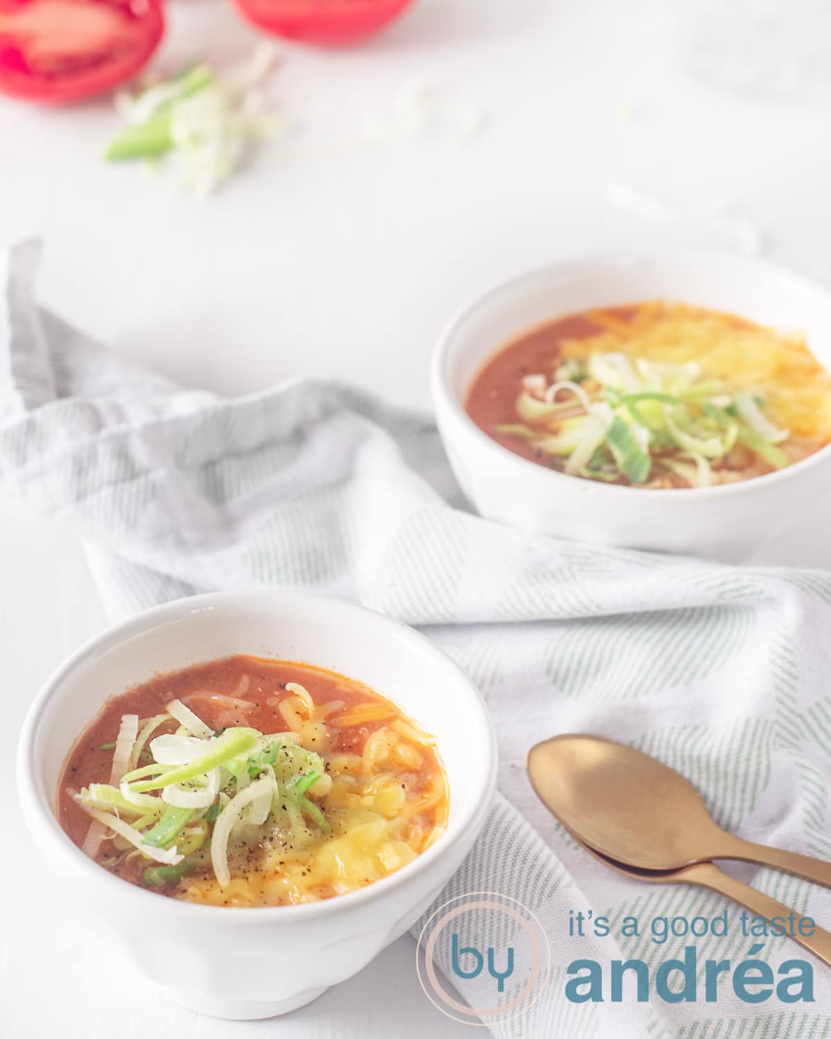 Two bowls filled with tomato soup with a cheese and leek topping. Two gold spoons on the right. A white green tea towel lies between the bowls.