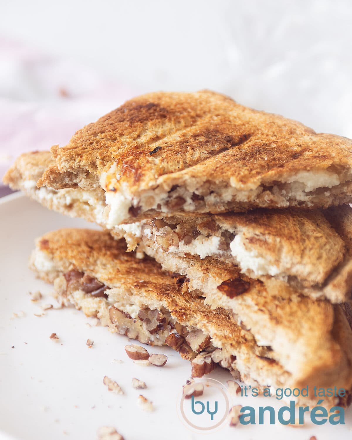 Een hoogtefoto met een macro van drie halve sneetjes tosti gevuld met geitenkaas, walnoten. In de achtergrond een witte ondergrond en een roze wit geblokte theedoek