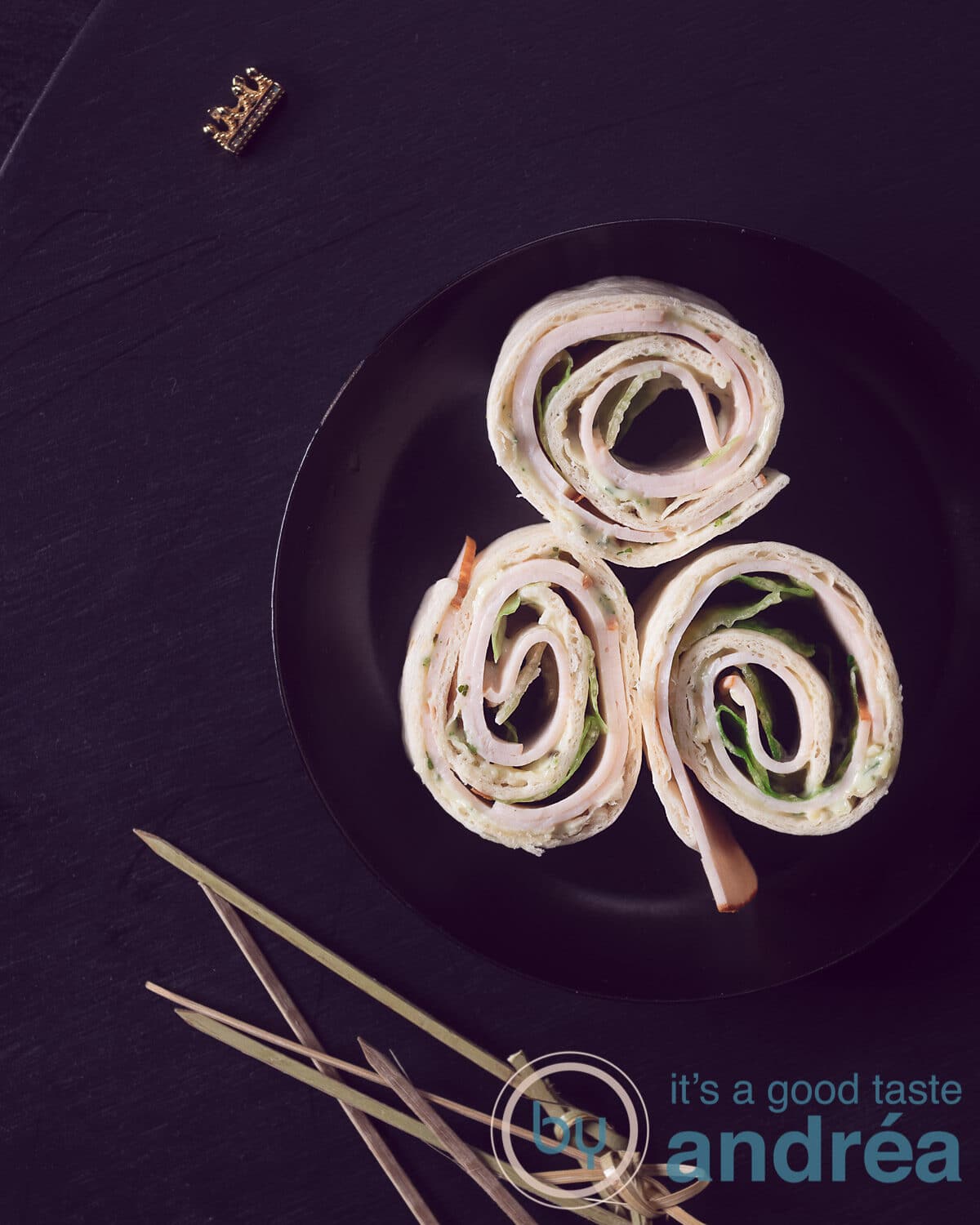 A photo from above with three wrap rolls with chicken, lettuce and witch's cheese on a black plate. Sticks at the bottom. Everything on a black background