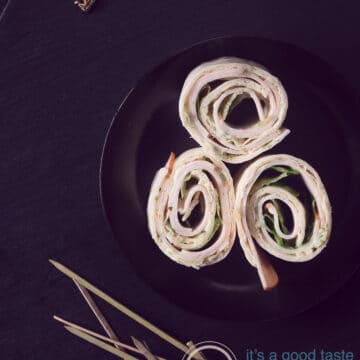 A photo from above with three wrap rolls with chicken, lettuce and witch's cheese on a black plate. Sticks at the bottom. Everything on a black background
