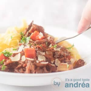 A square photo with a white plate filled with pappardelle and beef ragout with carrot and Parmesan cheese. A fork pierces it.