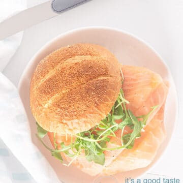 A photo from above of a sandwich filled with smoked salmon and arugula. At the top is a knife. Bottom left a white blue checkered tea towel.