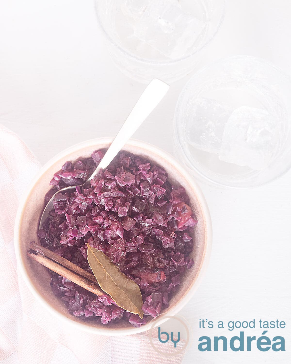 A photo from above of a pink dish filled with red cabbage with bay leaf and cinnamon on a white background. Two glasses at the top right