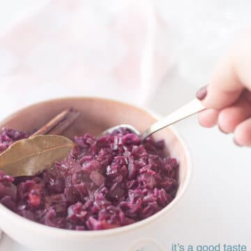 A photo of red cabbage in a pink dish with bay leaf and cinnamon on it. A hand takes a spoonful of red cabbage