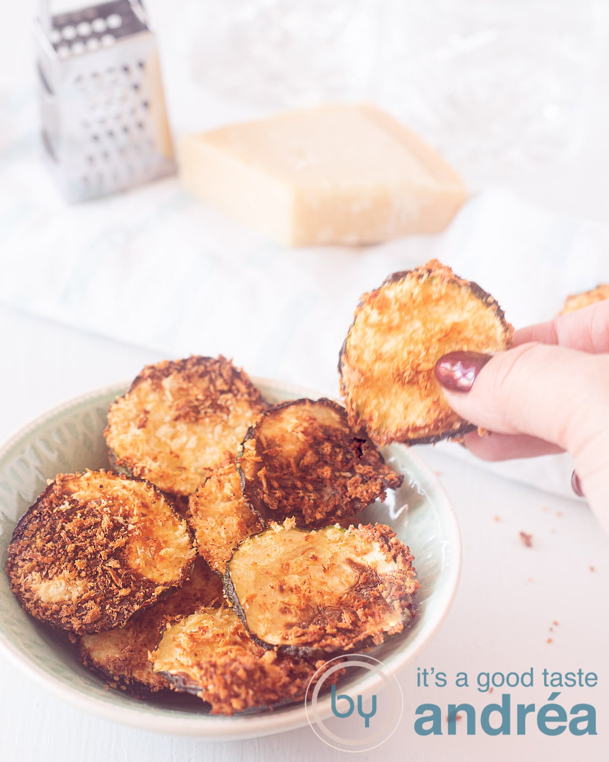 A hand taking a zucchini chips from a container full of chips. A white background. In the background of the photo Parmesan cheese and a grater.