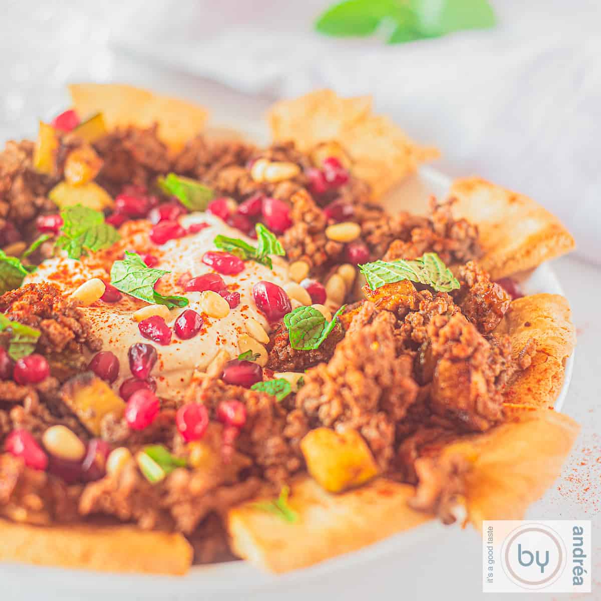 Een vierkante foto met een bord met pitapuntjes en fatteh met rundergehakt en courgette