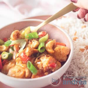 A pink bowl with chicken Siam and a gold spoon. Next to it is rice, and in the background is a pink and white tea towel.