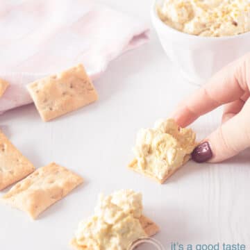 A hand grabbing a toast with chicken curry salad. A second piece of toast lies underneath. In the top left corner a pink white striped cloth and in the top right the rest of the salad
