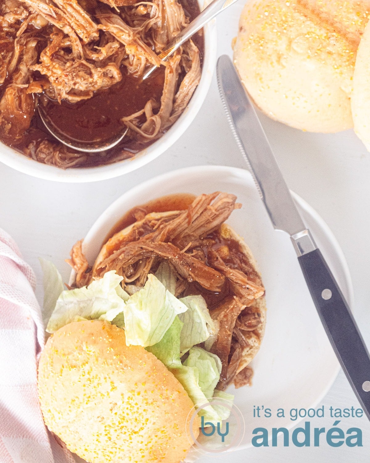 A photo from above of a pulled pork sandwich, extra sandwiches and a bowl of pulled pork on a white background
