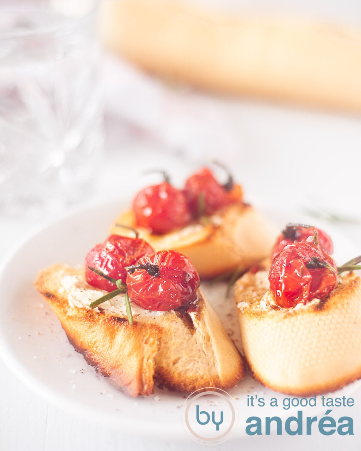An elevation photo with three grilled baguettes topped with goat cheese and stewed tomatoes on a white background. In the background a baguette and a glass.