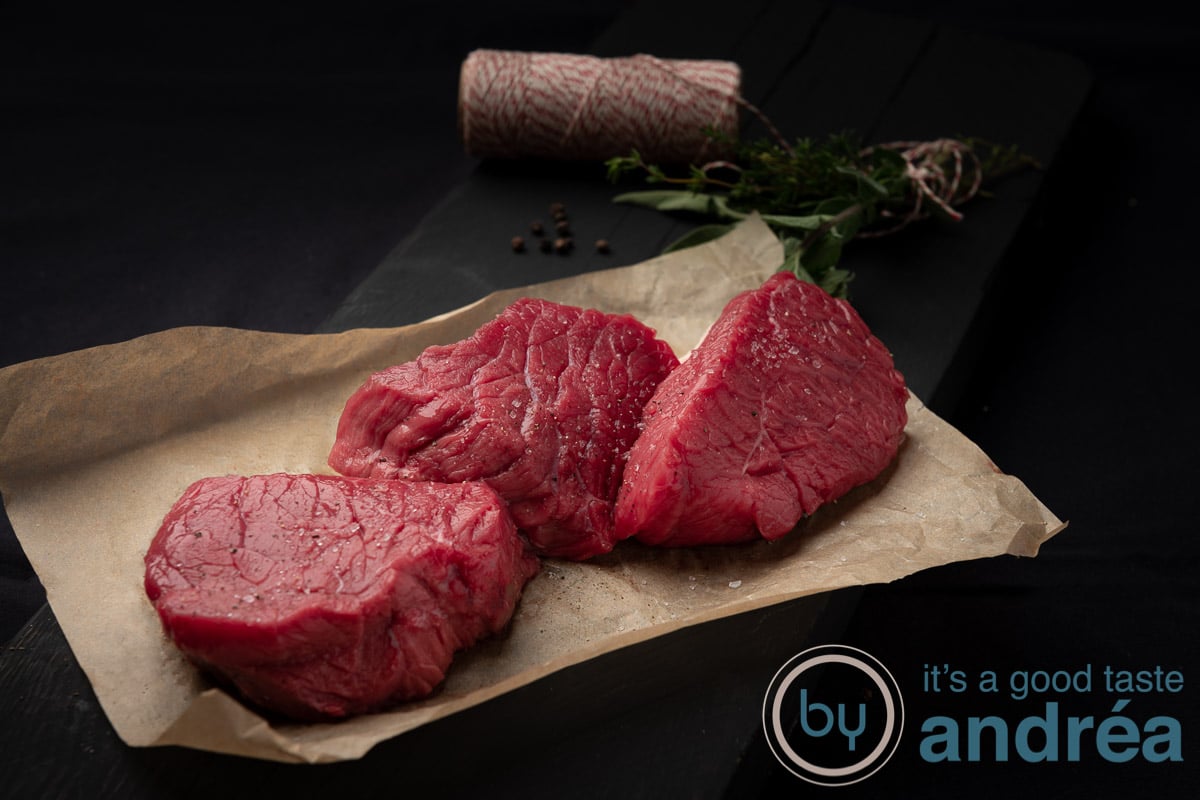 Three pieces of meat on a brown baking paper on a black background (steak) in the background twine and herbs