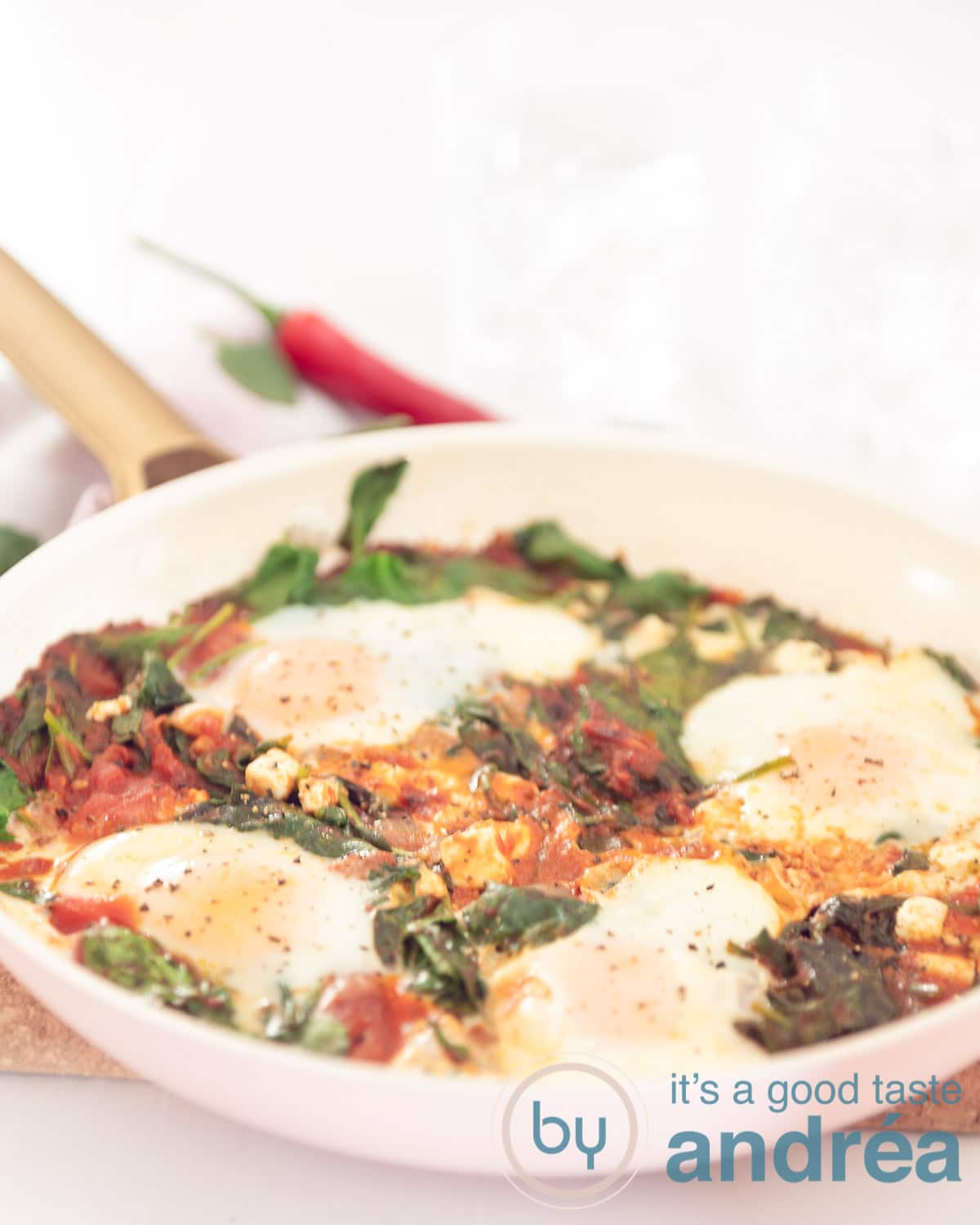 A pink pan filled with poached eggs in a tomato sauce, spinach and feta on a white background
