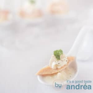 A square photo with a white spoon filled with asparagus mousse, trout and horseradish tuft on a white background in the background three more amuse spoons