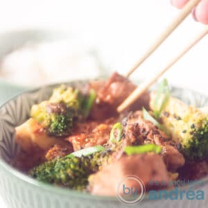 A square photo where two chopsticks take a cube of steak from a green bowl filled with broccoli, steak and sauce.
