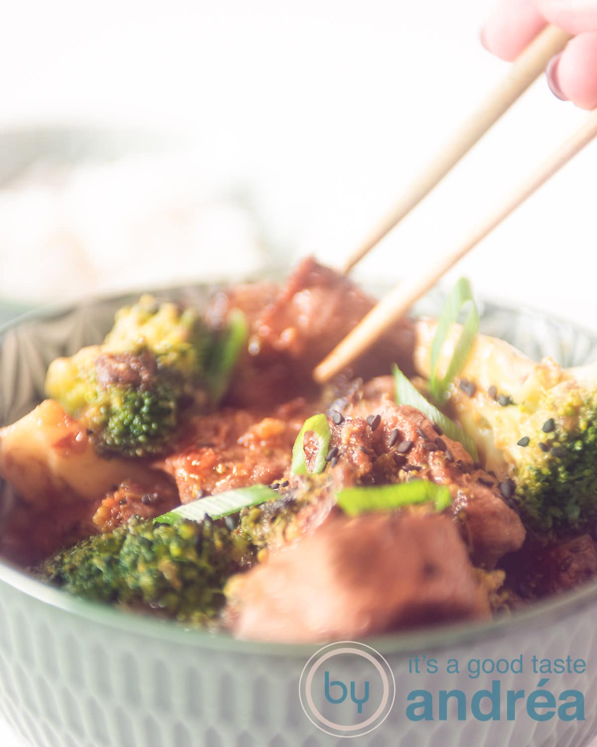 A bowl filled with steak, broccoli florets, mixed with a teriyaki sauce on a white background. Two chopsticks take a diced steak from the bowl.