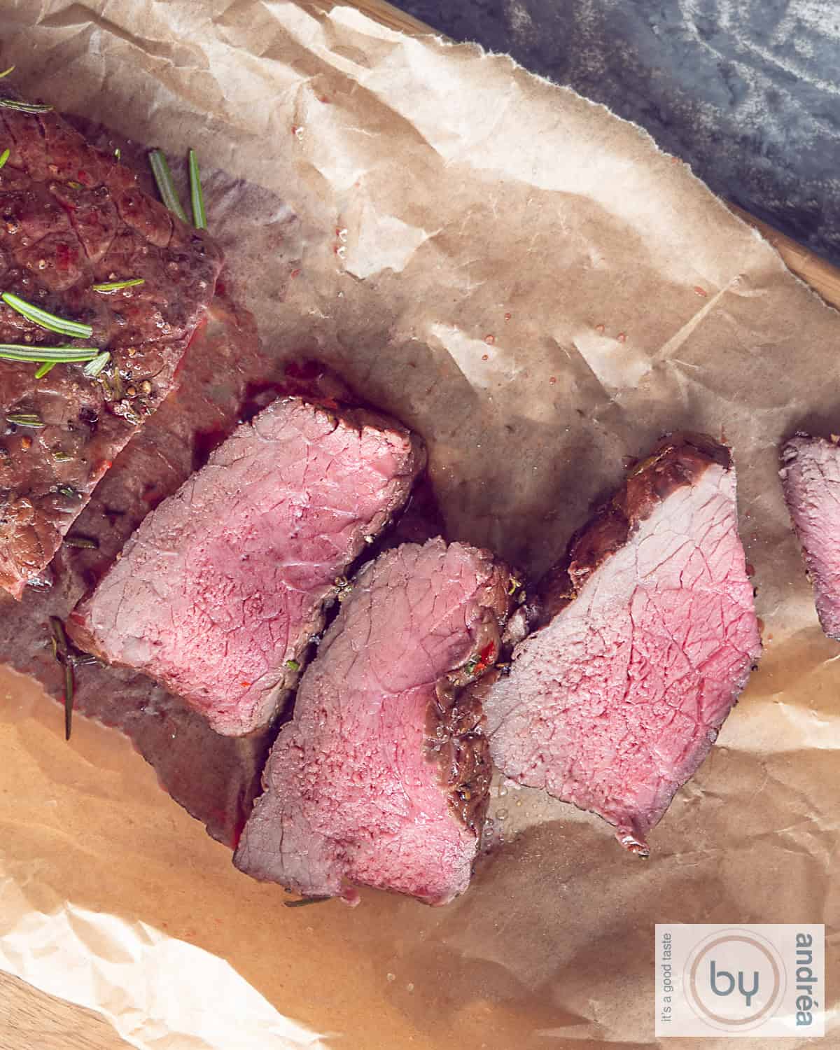 A photo from above of roast beef with three slices cut off on baking paper with fresh herbs.