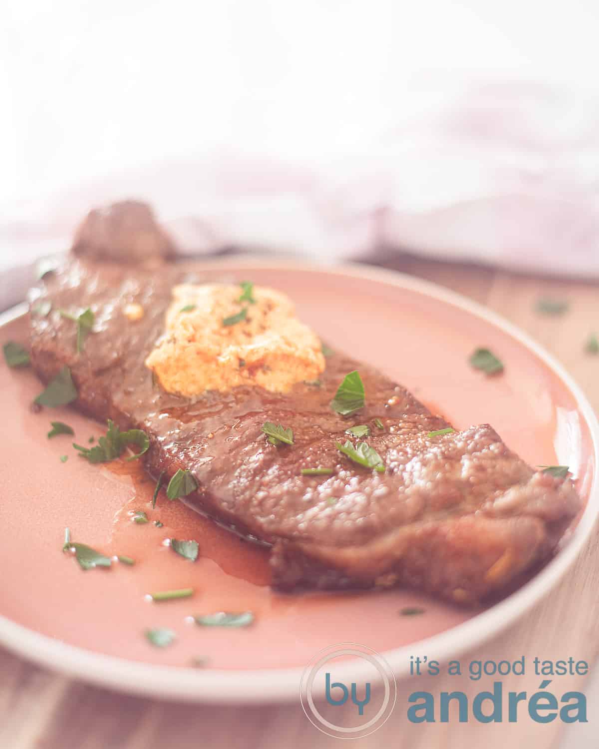 A pink plate on a wooden shelf with a pink and white checkered tea towel behind it. On the plate sirloin steak with herb butter and parsley