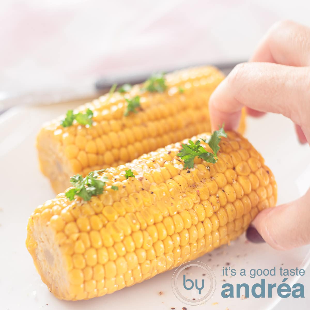 A square photo with two corncobs with parsley on a white background. A hand grabs the front corn cob.