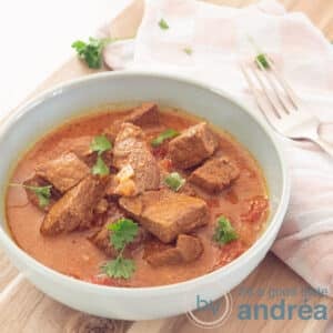 A square photo with a blue bowl filled with bombay curry on a wooden board