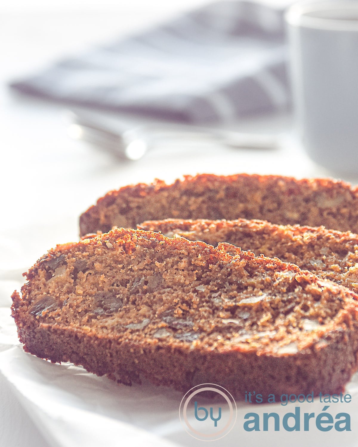 Three slices of date and apple cake on a white plate. A grey mug and cloth in the back.