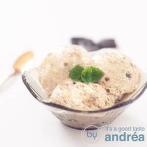 A square photo with a black bowl with three scoops of licorice ice cream, and a sprig of mint. Some liquorice and a golden spoon in the background