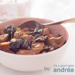 A square photo with a pink dish filled with beef stew, plums and carrot. A golden spoon sticks in it.