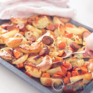 A square shot of a griddle filled with roast potato, carrot and smoked sausage. A spoon takes out a scoop. In the background a pink white tea towel.