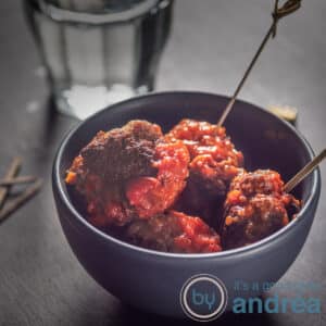 Een vierkante foto van een grijs bakje met gehaktballetjes in tomatensaus, prikkers erin gestoken.