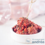 A square photograph of a white container with meatballs in tomato sauce, with a skewer inserted.