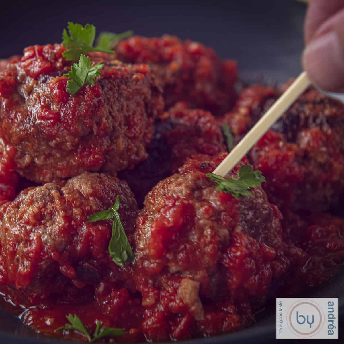 A square photo of meatballs in tomato sauce, Spanish. A skewer pierces a meatball.