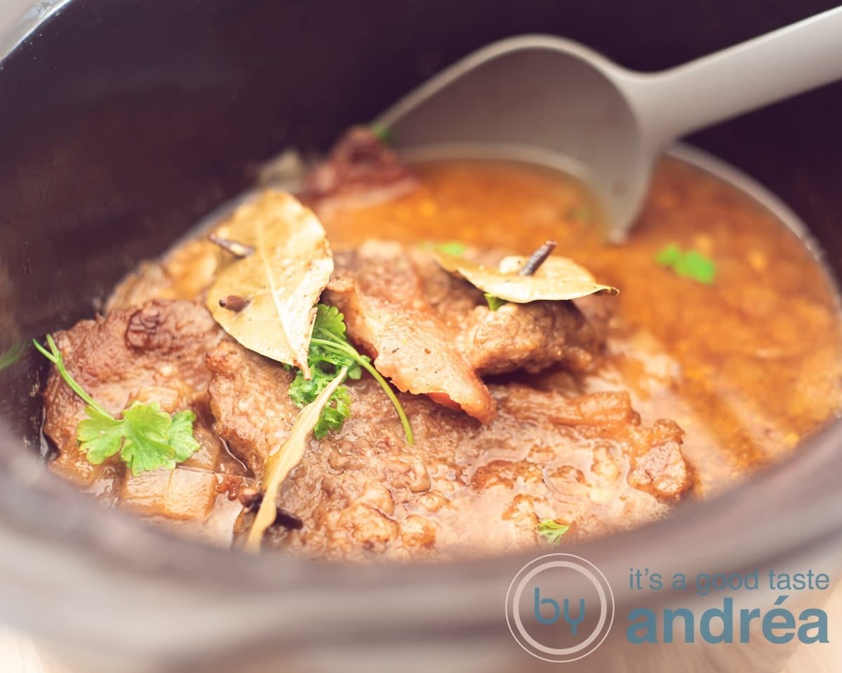 Een breedtefoto met een slowcooker gevuld met Stoofvlees, met laurierblad en peterselie. Een grijze opscheplepel neemt een beetje eruit.