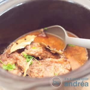 A square photo of a slow cooker filled with pot roast in a sauce. A gray spoon sticks into it.