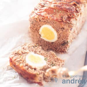 A square photo with meatloaf filled with egg and herbs on a baking paper. A golden fork takes a bite.