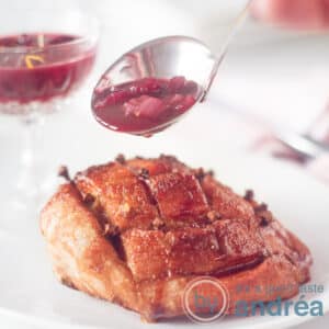 A square photo with a white plate of ham on the bone. A sauce ladle filled with cumberland sauce and some sauce in the background. A white background