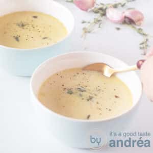 A square photo with two blue bowls filled with soup made from garlic, wine and broth. A hand takes a bite with a golden spoon. Garlic and thyme (fresh) in the background).