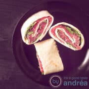 A square photo of a black background with a black plate with three tortilla snacks filled with carpaccio, arugula, parmesan cheese.
