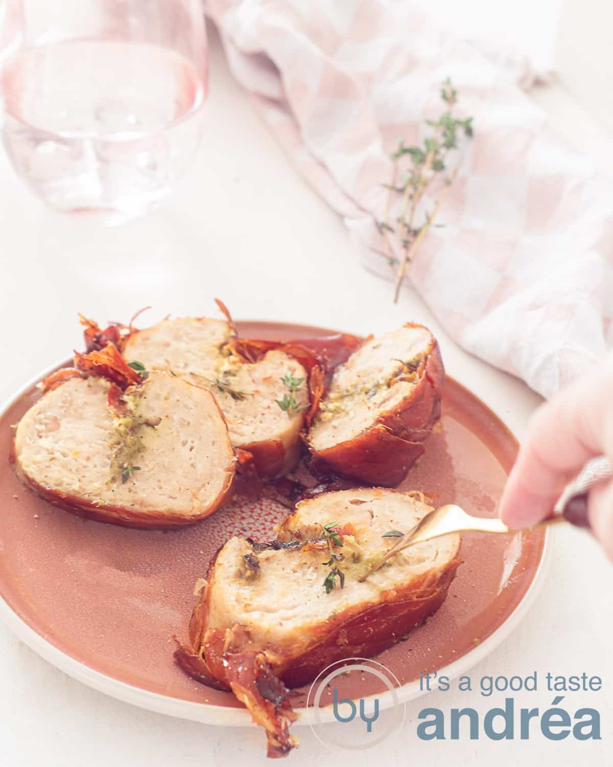 An elevation photo with a pink plate filled with chicken roulades and wrapped in prosciutto. A fork pierces it. A white background.