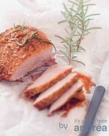 An elevation photo of a ham with maple glaze from which three slices have been cut off. Lying on a white baking paper, on the right side a steak knife and a rosemary sprig at the top