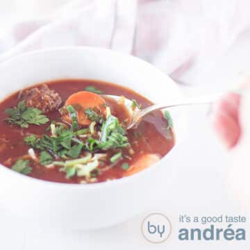 A square photo with a white bowl filled with carnival soup with sausage, leek and meatballs. A spoon takes a bite.