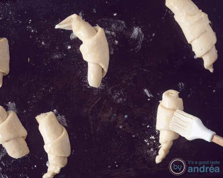 six croissants on a baking plate. A brush is giving an egg wash.