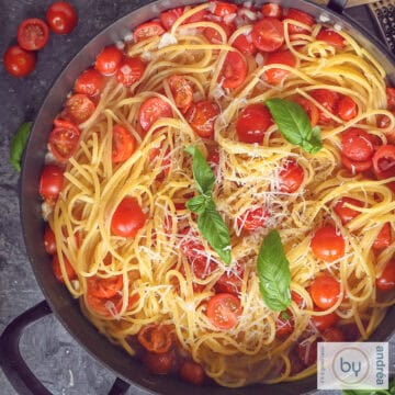 A square photo of a pink plate with linguine pasta, cherry tomatoes, basil, onion and cheese on a white background.