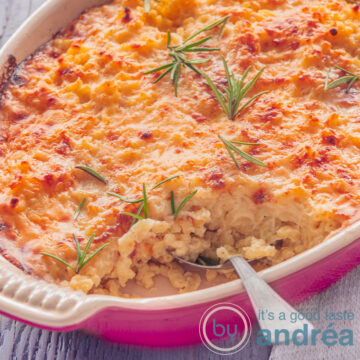 A square photo with part of a pink baking dish filled with macaroni and cheese, topped with sprigs of rosemary. A spoon sticks in. A gray background.