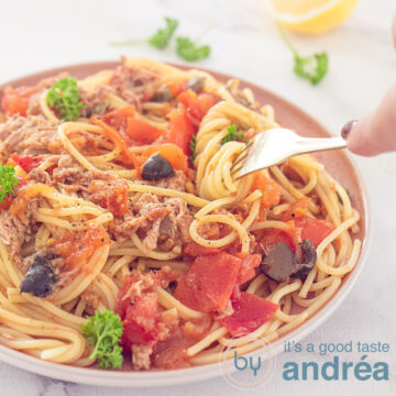A square photo with a pink plate with spaghetti Puttanesca on it. Spaghetti wrapped around a fork. A pink white tea towel in the background.