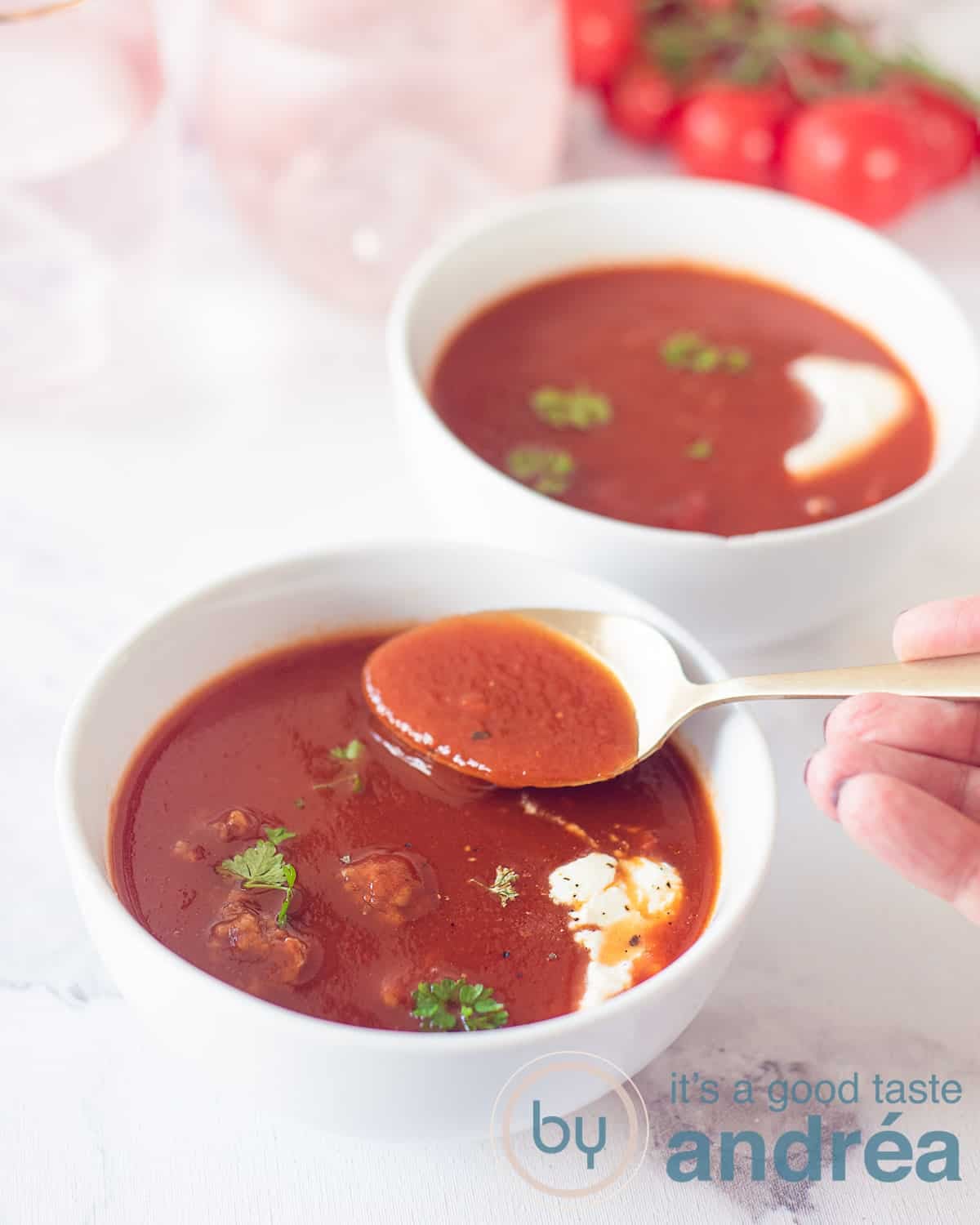 Two white bowls filled with tomato soup with meatballs and a dollop of cream. A hand with a spoon taking a bite.