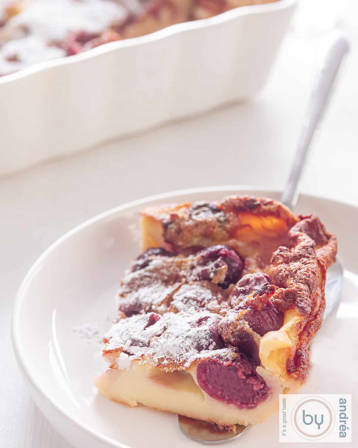 A piece of Cherry Clafoutis on a white plate. In the background a bowl with more cherry clafoutis