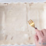 A white casserole dish covered with puff pastry. A fork pricking holes in the dough.