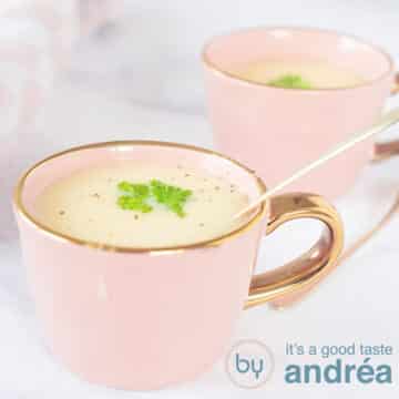 A square photo, two pink cups diagonally behind each other. In it a creamy asparagus soup with parsley. A white marbled background.