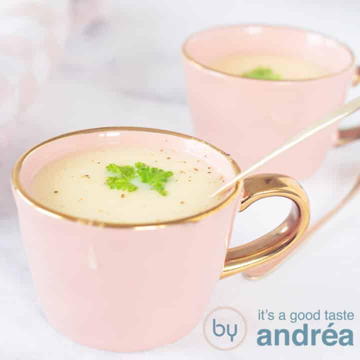 A square photo, two pink cups diagonally behind each other. In it a creamy asparagus soup with parsley. A white marbled background.