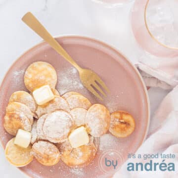 A square photo with a pink plate with a stack of mini pancakes, butter and icing sugar, a gold fork and a white background