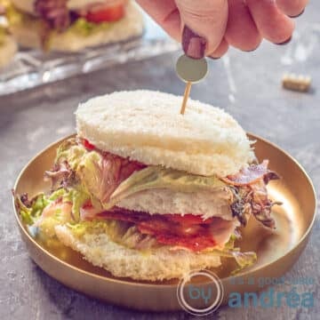 A square photo with a BLT mini sandwich on a gold plate on a gray background. A hand sticks a skewer in it.